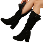 A pair of black knee-high slouchy boots with block heels and circular buckle details, shown on a model's legs against a white background.